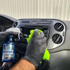 Ręce w czarnych rękawiczkach wykonują renowację plastików samochodowych, spryskując kokpit płynem Wash Mist i wycierając go zieloną mikrofibrą.