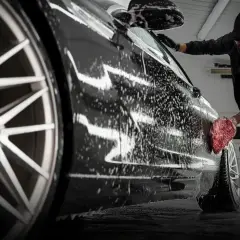 Profesjonalna konserwacja samochodu w pianie. Mężczyzna myje czarne auto gąbką, obok butelka szamponu SWAG.