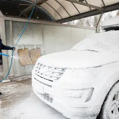 Mężczyzna w zimowej kurtce i czapce myje białe auto na myjni. Czy można myć auto w zimie? Tak, jeśli używasz odpowiednich środków.