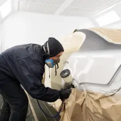 Lakiernik w masce i kombinezonie maluje samochód w kabinie. Czas, ile schnie lakier samochodowy, zależy od wielu czynników.