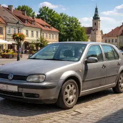 Szary Volkswagen Golf IV na brukowanym rynku. Wypłowiały kolor lakieru świadczy o latach służby. W tle kamienice i wieża kościelna.