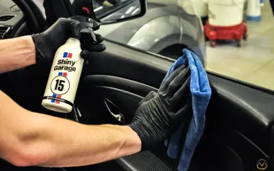 Ręce w czarnych rękawiczkach używają preparatu Shiny Garage Apple Dressing i niebieskiej mikrofibry, by dowiedzieć się, czym wyczyścić plastiki w samochodzie.