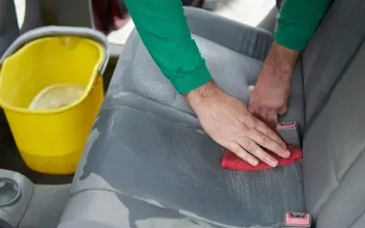 Czyszczenie tapicerki samochodowej z plam po oleju silnikowym. Ręce wycierają siedzenie czerwoną gąbką, obok stoi żółte wiadro z wodą.