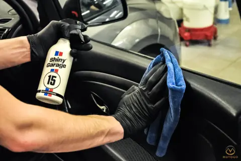 Ręce w czarnych rękawiczkach używają preparatu Shiny Garage Apple Dressing i niebieskiej mikrofibry, by dowiedzieć się, czym wyczyścić plastiki w samochodzie.