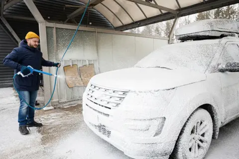 Mężczyzna w zimowej kurtce i czapce myje białe auto na myjni. Czy można myć auto w zimie? Tak, jeśli używasz odpowiednich środków.