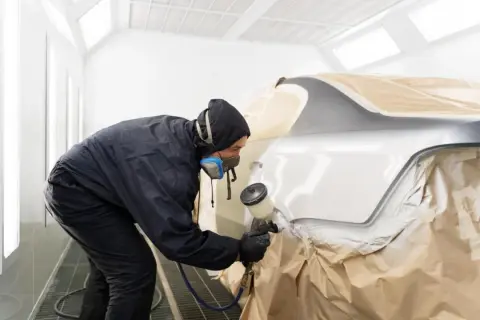 Lakiernik w masce i kombinezonie maluje samochód w kabinie. Czas, ile schnie lakier samochodowy, zależy od wielu czynników.