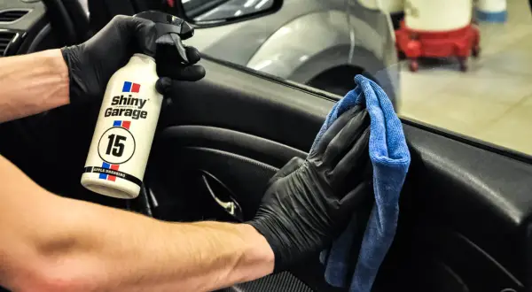 Ręce w czarnych rękawiczkach używają preparatu Shiny Garage Apple Dressing i niebieskiej mikrofibry, by dowiedzieć się, czym wyczyścić plastiki w samochodzie.