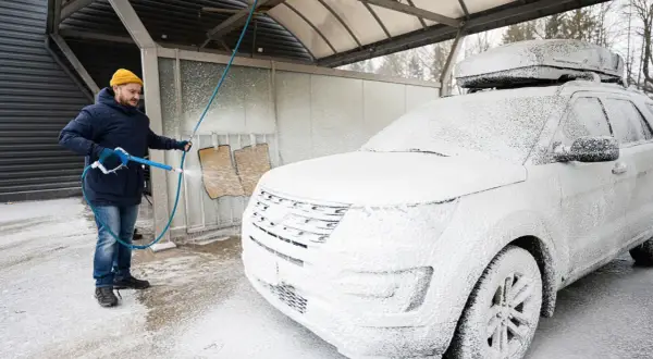 Mężczyzna w zimowej kurtce i czapce myje białe auto na myjni. Czy można myć auto w zimie? Tak, jeśli używasz odpowiednich środków.