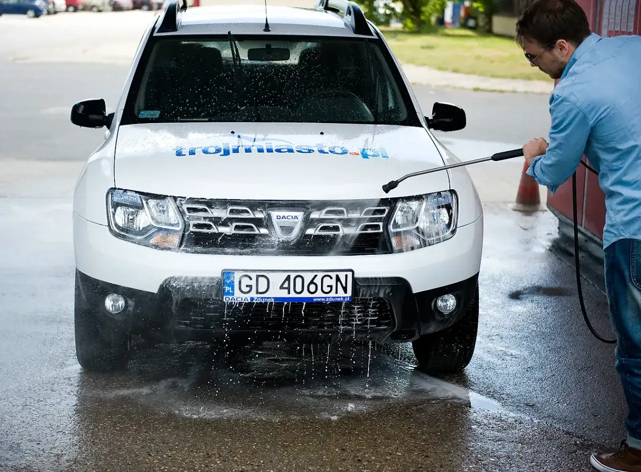 Mężczyzna myje białe Dacia na myjni bezdotykowej. Woda spływa po masce, a na szybie widać krople. Poznaj myjnia bezdotykowa triki!
