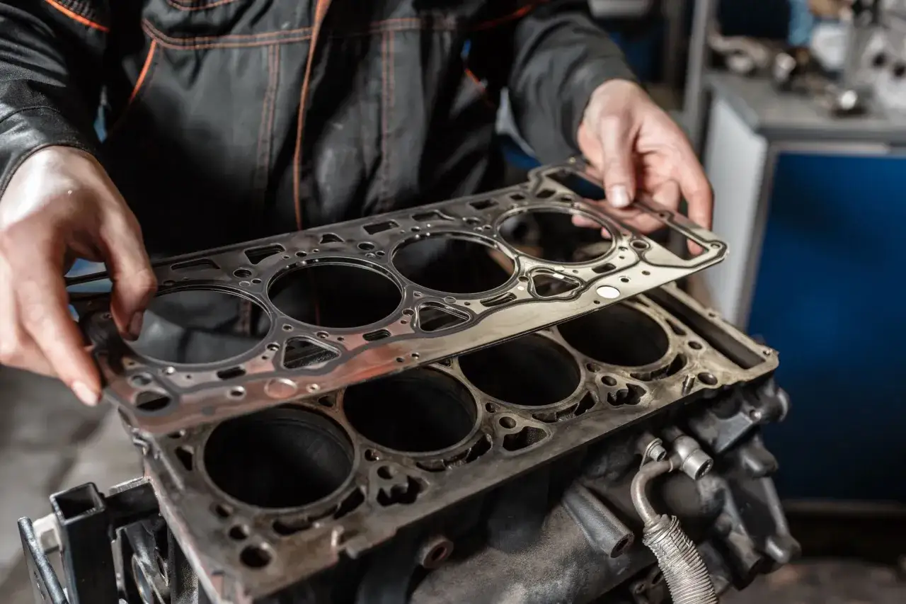 Mechanik przygotowuje wymianę uszczelek w samochodzie, trzymając nową uszczelkę nad blokiem silnika.