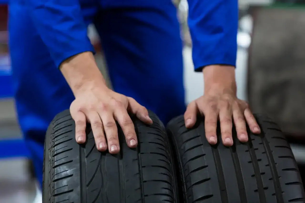 Dłonie mechanika w niebieskim kombinezonie dotykają dw&oacute;ch opon. Jedna z nich ma wyraźnie wyząbkowane opony, co sugeruje zużycie.