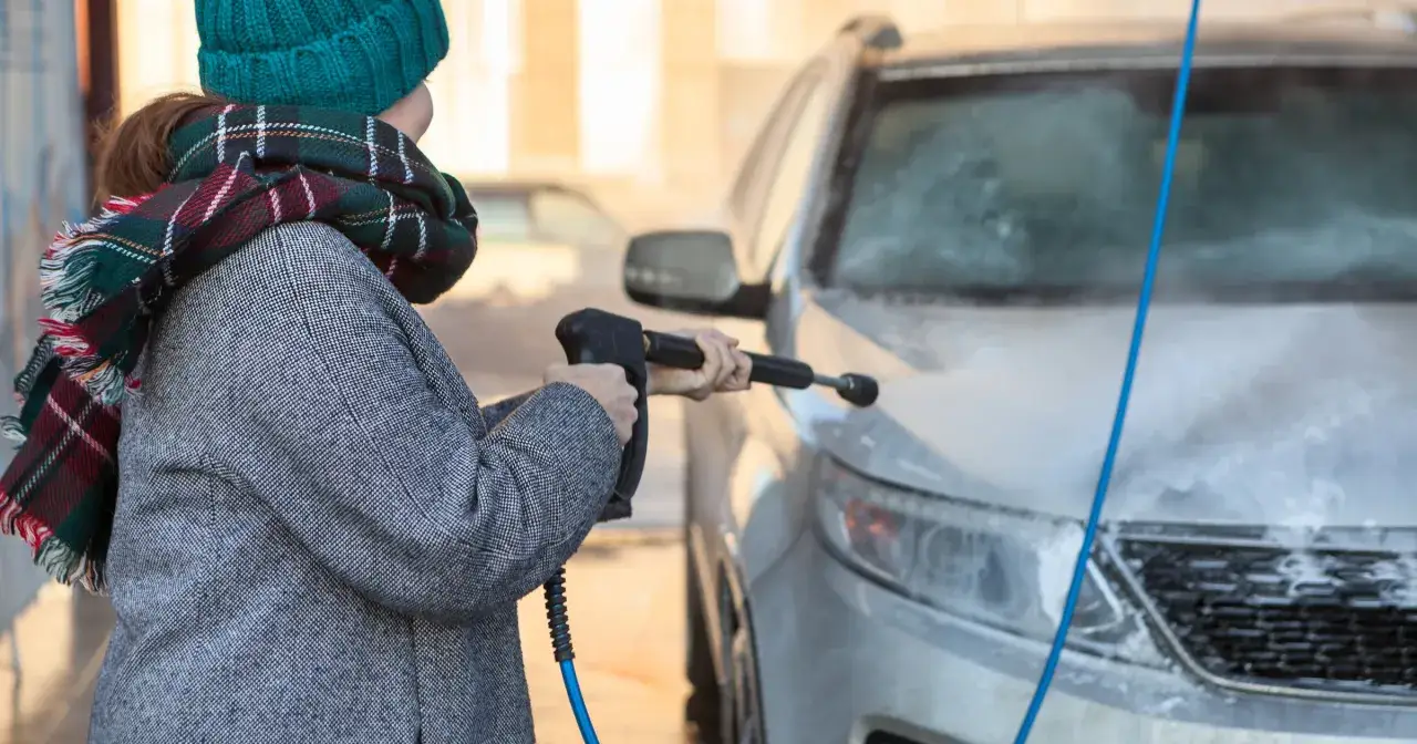 Kobieta w czapce i szaliku myje auto zimą na myjni bezdotykowej, używając myjki ciśnieniowej.
